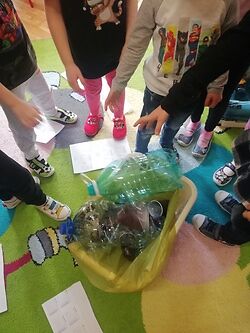 Zdjęcie przestawia dzieci przedszkolne segregujące odpady plastiku, w tym butelki plastikowe do żółtego worka, w trakcie warsztatów edukacyjnych, nie są widoczne twarze dzieci, zdjecie jest wykonane w kolorze.